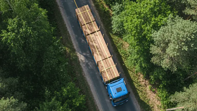 Aerial view of a blue truck transporting wooden boards along a road surrounded by forest.